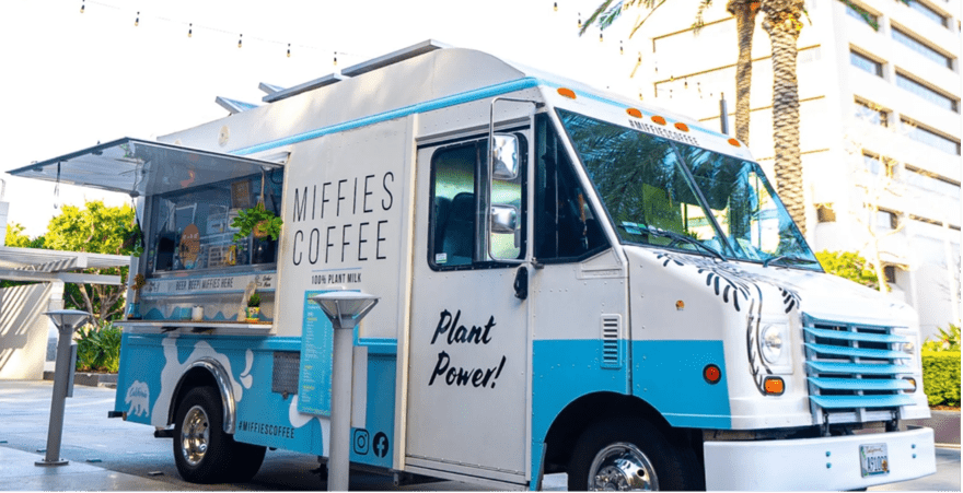 A white food truck named Miffie's Coffee is parked in front of a tall building. The truck sells 100% plant-based coffee and other drinks.